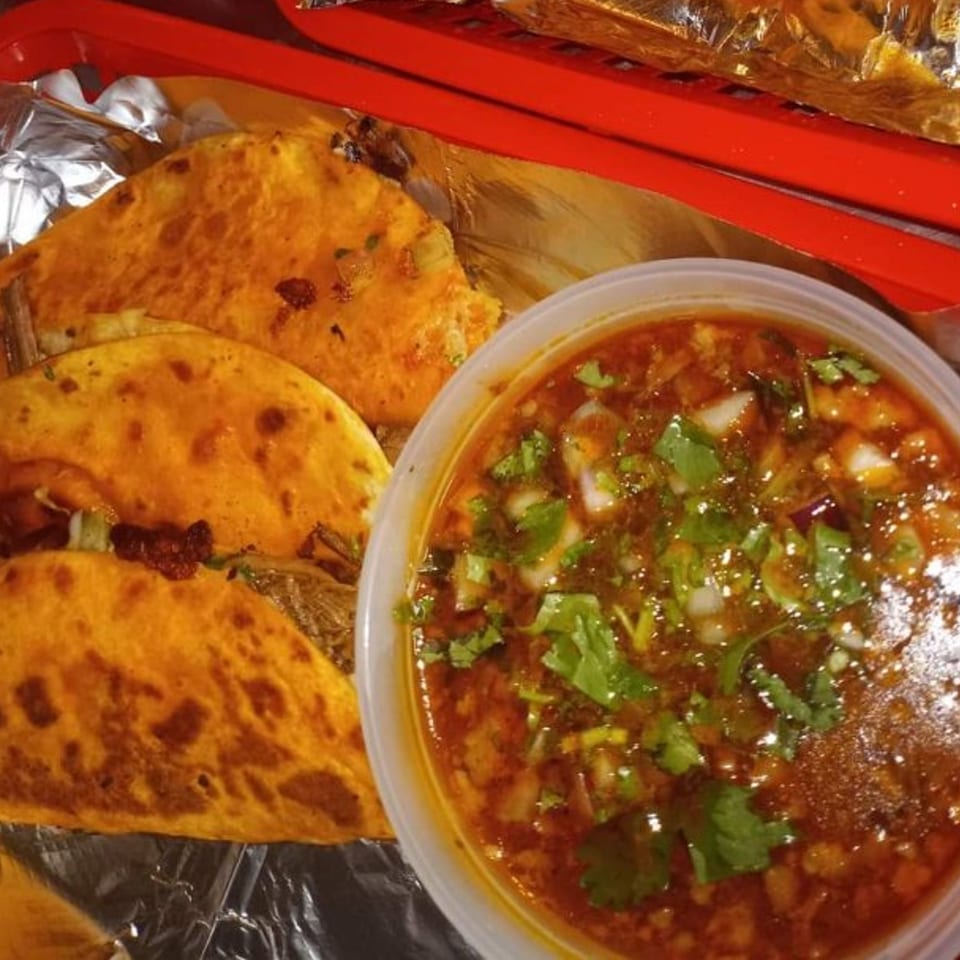 Three Sazón Bello Taquería birria tacos with a side of consommé, garnished with cilantro and onions.