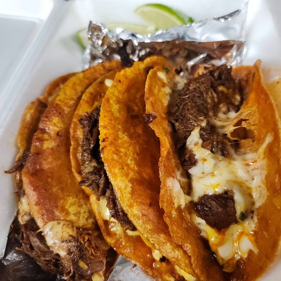 Four cheesy birria tacos with shredded beef and lime wedges in a white takeout container.