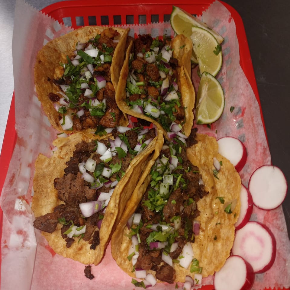 Four Sazón Bello Taquería tacos with meat, onions, and cilantro, served with lime and radish slices.